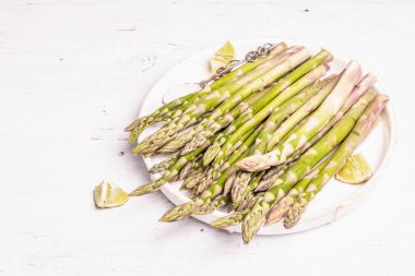 Olgun kuşkonmaz pişirmeye hazır. Seramik tabak ve limon parçaları. Modern sert ışık, karanlık gölge. Beyaz tahta masa, boşluğu kopyala