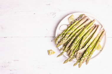 Olgun kuşkonmaz pişirmeye hazır. Seramik tabak ve limon parçaları. Modern sert ışık, karanlık gölge. Beyaz ahşap masa, üst manzara.