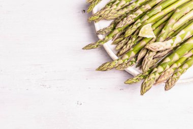 Olgun kuşkonmaz pişirmeye hazır. Seramik tabak ve limon parçaları. Modern sert ışık, karanlık gölge. Beyaz ahşap masa, üst manzara.