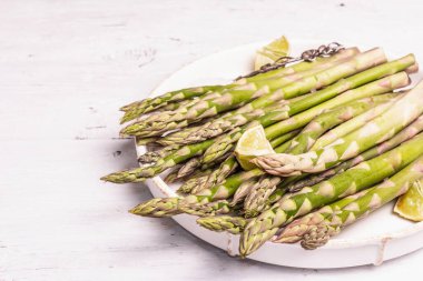 Olgun kuşkonmaz pişirmeye hazır. Seramik tabak ve limon parçaları. Modern sert ışık, karanlık gölge. Beyaz tahta masa, boşluğu kopyala