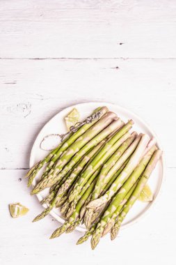 Olgun kuşkonmaz pişirmeye hazır. Seramik tabak ve limon parçaları. Modern sert ışık, karanlık gölge. Beyaz ahşap masa, üst manzara.