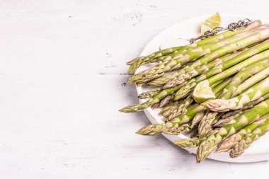 Olgun kuşkonmaz pişirmeye hazır. Seramik tabak ve limon parçaları. Modern sert ışık, karanlık gölge. Beyaz tahta masa, boşluğu kopyala