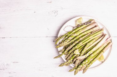 Olgun kuşkonmaz pişirmeye hazır. Seramik tabak ve limon parçaları. Modern sert ışık, karanlık gölge. Beyaz ahşap masa, üst manzara.