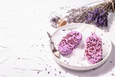 Mor Lavanta aromalı dondurma. Çiçek buketi, klasik gümüş kaşık, seramik standı. Modern sert ışık, karanlık gölge. Beyaz macun arkaplanı, kopyalama alanı