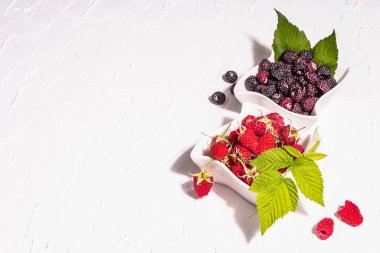 Çeşit çeşit kırmızı ve siyah ahududu. Seramik kaseler, modern sert ışık, karanlık gölge. Beyaz mutfak arka planı, kapat