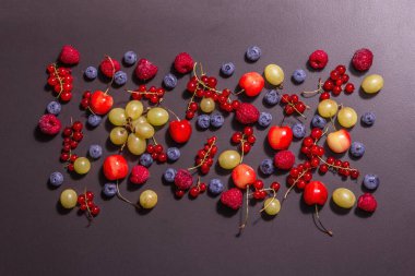 Değişik olgun meyveler, siyah bir taşın üzerine serpiştirilmiş. Taze ahududu, yabanmersini, üzüm, tatlı kiraz ve kırmızı frenk üzümü. Modern sert bir ışık, karanlık bir gölge, üst manzara.
