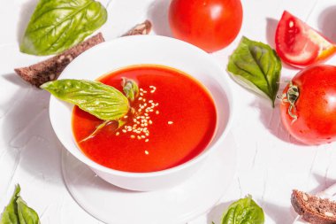 Ev yapımı domates çorbası dolu kâseler. Olgun sebzeler, taze fesleğen, ekmek çubukları, aromatik baharatlar. Modaya uygun sert ışık, karanlık gölge. Beyaz macun arkaplan, düz yatar