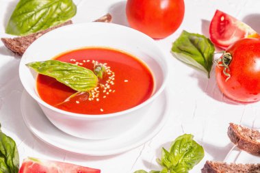 Ev yapımı domates çorbası dolu kâseler. Olgun sebzeler, taze fesleğen, ekmek çubukları, aromatik baharatlar. Modaya uygun sert ışık, karanlık gölge. Beyaz macun arkaplan, düz yatar