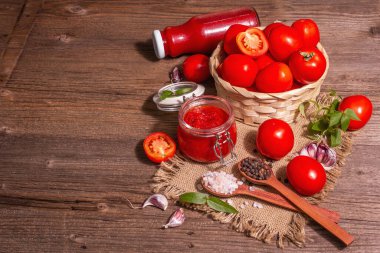 Domates şenliği, reçel, turşu, cam kavanozdaki sos. Ev yapımı koruma konsepti. Taze domates, baharat, sarımsak, yeşillik, deniz tuzu. Modern sert ışık, karanlık gölge, eski ahşap masa, kopyalama alanı