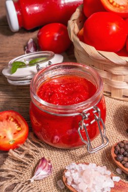 Domates şenliği, reçel, turşu, cam kavanozdaki sos. Ev yapımı koruma konsepti. Taze domates, baharat, sarımsak, yeşillik, deniz tuzu. Modern sert bir ışık, karanlık bir gölge, eski bir ahşap masa, yakın çekim.