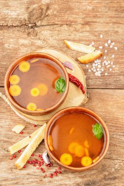 Seramik kaselerde sebzeli şeffaf tavuk suyu çorbası. Geleneksel bulyon, sağlıklı yemek, çatal bıçak takımı. Eski ahşap zemin, üst manzara