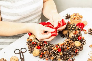 Dişi eller sonbahar koni, fındık ve böğürtlen çelengi için bağlı bir yay tutar. Yaratıcı kompozisyon için doğal malzemeler. El yapımı ev dekorasyonu şenlik konsepti. Beyaz ahşap masa