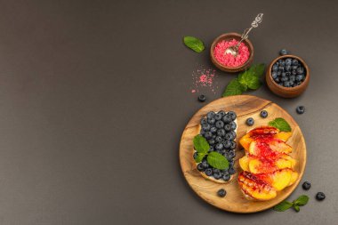 Krem peynirli ve olgun meyveli tost ekmeği. Izgara şeftali, yabanmersini ve siyah beton arka planda nane yaprakları. Yararlı kahvaltı konsepti, üst görünüm