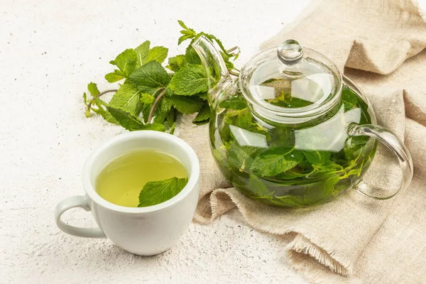 Fragrant mint tea on a light background. Glass teapot, a cup of tea, a bunch of fresh mint. Good morning concept, close up