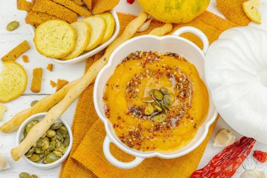 Tohumlar ve baharatlarla süslenmiş bir kase balkabağı humusu, Grissini ve kumaş peçetede kızarmış ekmeklerle servis ediliyor. Beyaz ahşap tahtalar, vejetaryen sağlıklı gıda konsepti, üst manzara.