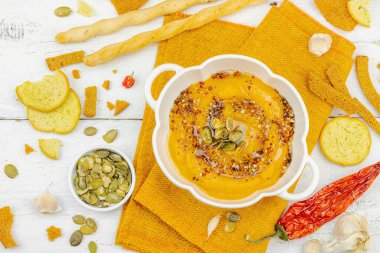 Tohumlar ve baharatlarla süslenmiş bir kase balkabağı humusu, Grissini ve kumaş peçetede kızarmış ekmeklerle servis ediliyor. Beyaz ahşap tahtalar, vejetaryen sağlıklı gıda konsepti, üst manzara.