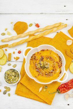 Tohumlar ve baharatlarla süslenmiş bir kase balkabağı humusu, Grissini ve kumaş peçetede kızarmış ekmeklerle servis ediliyor. Beyaz ahşap tahtalar, vejetaryen sağlıklı gıda konsepti, üst manzara.