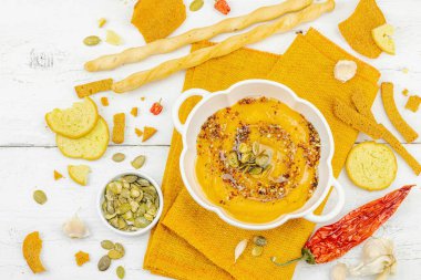 Tohumlar ve baharatlarla süslenmiş bir kase balkabağı humusu, Grissini ve kumaş peçetede kızarmış ekmeklerle servis ediliyor. Beyaz ahşap tahtalar, vejetaryen sağlıklı gıda konsepti, üst manzara.