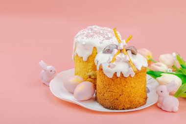 Geleneksel Paskalya pastaları, beyaz kremalı ve tabakta renkli yumurtalar serpiştirilmiş. Pembe arka planda tavşanları ve laleleri olan şenlikli bir kompozisyon. Ev yapımı tatil pişirme konsepti, kopyalama alanı