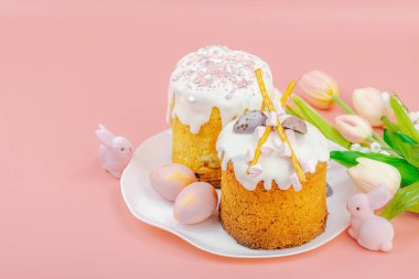 Geleneksel Paskalya pastaları, beyaz kremalı ve tabakta renkli yumurtalar serpiştirilmiş. Pembe arka planda tavşanları ve laleleri olan şenlikli bir kompozisyon. Ev yapımı tatil pişirme konsepti, kopyalama alanı