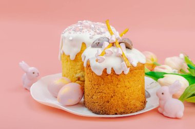 Geleneksel Paskalya pastaları, beyaz kremalı ve tabakta renkli yumurtalar serpiştirilmiş. Pembe arka planda tavşanları ve laleleri olan şenlikli bir kompozisyon. Ev yapımı tatil pişirme konsepti, kopyalama alanı