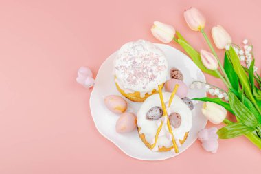 Geleneksel Paskalya pastaları, beyaz kremalı ve tabakta renkli yumurtalar serpiştirilmiş. Pembe arka planda tavşanları ve laleleri olan şenlikli bir kompozisyon. Ev yapımı tatil pişirme konsepti, üst görünüm