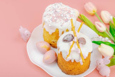 Geleneksel Paskalya pastaları, beyaz kremalı ve tabakta renkli yumurtalar serpiştirilmiş. Pembe arka planda tavşanları ve laleleri olan şenlikli bir kompozisyon. Ev yapımı tatil pişirme konsepti, kopyalama alanı