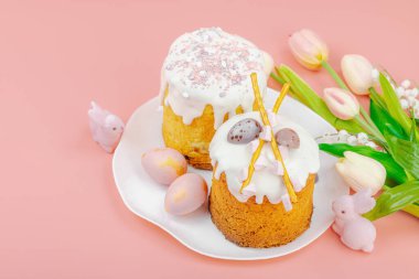 Geleneksel Paskalya pastaları, beyaz kremalı ve tabakta renkli yumurtalar serpiştirilmiş. Pembe arka planda tavşanları ve laleleri olan şenlikli bir kompozisyon. Ev yapımı tatil pişirme konsepti, kopyalama alanı