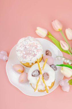 Geleneksel Paskalya pastaları, beyaz kremalı ve tabakta renkli yumurtalar serpiştirilmiş. Pembe arka planda tavşanları ve laleleri olan şenlikli bir kompozisyon. Ev yapımı tatil pişirme konsepti, üst görünüm