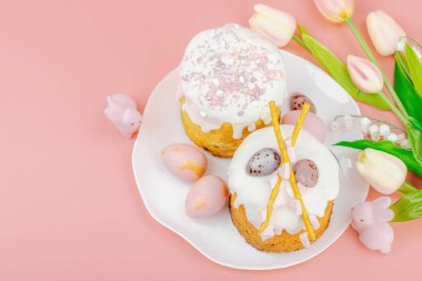 Geleneksel Paskalya pastaları, beyaz kremalı ve tabakta renkli yumurtalar serpiştirilmiş. Pembe arka planda tavşanları ve laleleri olan şenlikli bir kompozisyon. Ev yapımı tatil pişirme konsepti, üst görünüm