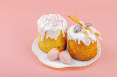 Geleneksel Paskalya pastaları, beyaz kremalı ve tabakta renkli yumurtalar serpiştirilmiş. Pembe arka planda tavşanları ve laleleri olan şenlikli bir kompozisyon. Ev yapımı tatil pişirme konsepti, kopyalama alanı