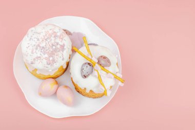 Geleneksel Paskalya pastaları, beyaz kremalı ve tabakta renkli yumurtalar serpiştirilmiş. Pembe arka planda tavşanları ve laleleri olan şenlikli bir kompozisyon. Ev yapımı tatil pişirme konsepti, üst görünüm