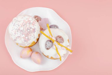 Geleneksel Paskalya pastaları, beyaz kremalı ve tabakta renkli yumurtalar serpiştirilmiş. Pembe arka planda tavşanları ve laleleri olan şenlikli bir kompozisyon. Ev yapımı tatil pişirme konsepti, üst görünüm