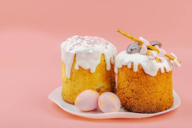 Geleneksel Paskalya pastaları, beyaz kremalı ve tabakta renkli yumurtalar serpiştirilmiş. Pembe arka planda tavşanları ve laleleri olan şenlikli bir kompozisyon. Ev yapımı tatil pişirme konsepti, kopyalama alanı