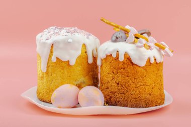 Geleneksel Paskalya pastaları, beyaz kremalı ve tabakta renkli yumurtalar serpiştirilmiş. Pembe arka planda tavşanları ve laleleri olan şenlikli bir kompozisyon. Ev yapımı tatil pişirme konsepti. Kapatın.