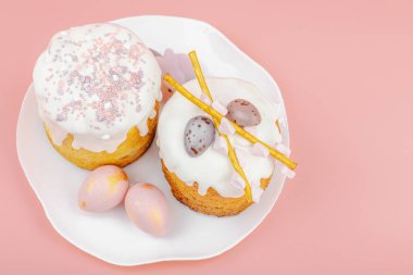 Geleneksel Paskalya pastaları, beyaz kremalı ve tabakta renkli yumurtalar serpiştirilmiş. Pembe arka planda tavşanları ve laleleri olan şenlikli bir kompozisyon. Ev yapımı tatil pişirme konsepti, üst görünüm