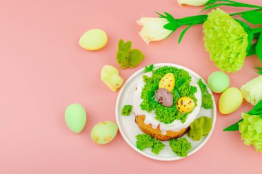 Geleneksel Paskalya pastaları, beyaz kremalı ve tabakta renkli yumurtalar serpiştirilmiş. Pembe arka planda tavşanlı ve çiçekli şenlikli kompozisyon. Ev yapımı tatil pişirme konsepti, üst görünüm