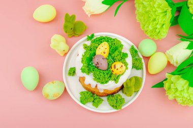 Geleneksel Paskalya pastaları, beyaz kremalı ve tabakta renkli yumurtalar serpiştirilmiş. Pembe arka planda tavşanlı ve çiçekli şenlikli kompozisyon. Ev yapımı tatil pişirme konsepti, üst görünüm