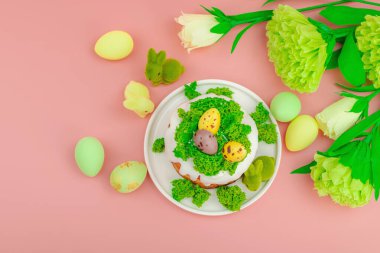 Geleneksel Paskalya pastaları, beyaz kremalı ve tabakta renkli yumurtalar serpiştirilmiş. Pembe arka planda tavşanlı ve çiçekli şenlikli kompozisyon. Ev yapımı tatil pişirme konsepti, üst görünüm