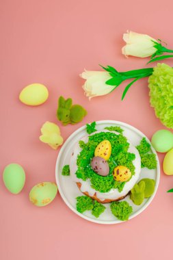 Geleneksel Paskalya pastaları, beyaz kremalı ve tabakta renkli yumurtalar serpiştirilmiş. Pembe arka planda tavşanlı ve çiçekli şenlikli kompozisyon. Ev yapımı tatil pişirme konsepti, üst görünüm