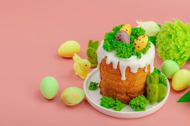 Geleneksel Paskalya pastaları, beyaz kremalı ve tabakta renkli yumurtalar serpiştirilmiş. Pembe arka planda tavşanlı ve çiçekli şenlikli kompozisyon. Ev yapımı tatil pişirme konsepti, kopyalama alanı