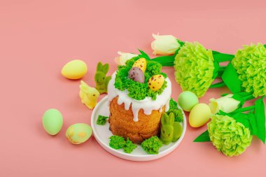 Geleneksel Paskalya pastaları, beyaz kremalı ve tabakta renkli yumurtalar serpiştirilmiş. Pembe arka planda tavşanlı ve çiçekli şenlikli kompozisyon. Ev yapımı tatil pişirme konsepti, kopyalama alanı
