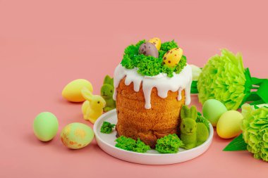 Geleneksel Paskalya pastaları, beyaz kremalı ve tabakta renkli yumurtalar serpiştirilmiş. Pembe arka planda tavşanlı ve çiçekli şenlikli kompozisyon. Ev yapımı tatil pişirme konsepti, kopyalama alanı