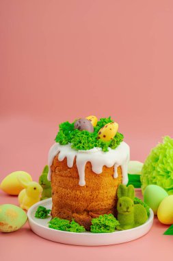 Geleneksel Paskalya pastaları, beyaz kremalı ve tabakta renkli yumurtalar serpiştirilmiş. Pembe arka planda tavşanlı ve çiçekli şenlikli kompozisyon. Ev yapımı tatil pişirme konsepti, kopyalama alanı
