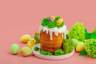 Geleneksel Paskalya pastaları, beyaz kremalı ve tabakta renkli yumurtalar serpiştirilmiş. Pembe arka planda tavşanlı ve çiçekli şenlikli kompozisyon. Ev yapımı tatil pişirme konsepti, kopyalama alanı