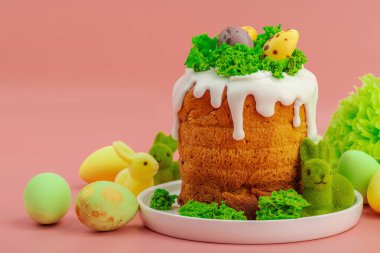 Geleneksel Paskalya pastaları, beyaz kremalı ve tabakta renkli yumurtalar serpiştirilmiş. Pembe arka planda tavşanlı ve çiçekli şenlikli kompozisyon. Ev yapımı tatil pişirme konsepti. Kapatın.