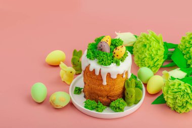 Geleneksel Paskalya pastaları, beyaz kremalı ve tabakta renkli yumurtalar serpiştirilmiş. Pembe arka planda tavşanlı ve çiçekli şenlikli kompozisyon. Ev yapımı tatil pişirme konsepti, kopyalama alanı