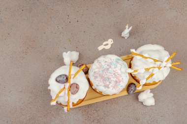 Traditional Easter cakes with white frosting and decorative pussy willow branches on wooden stand. Festive still life with pastel eggs and bunny figures on stone background, top view
