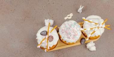 Traditional Easter cakes with white frosting and decorative pussy willow branches on wooden stand. Festive still life with pastel eggs and bunny figures on stone background, banner format