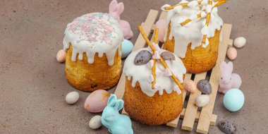 Traditional Easter cakes with white frosting and decorative pussy willow branches on wooden stand. Festive still life with pastel eggs and bunny figures on stone background, banner format